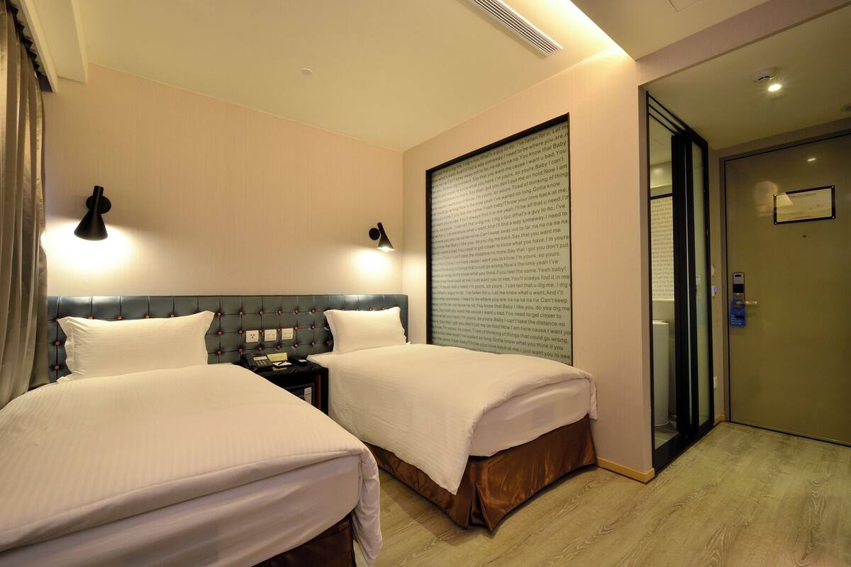 Habitación de hotel con dos camas, una mesita de noche, una lámpara de pared y una puerta con panel de vidrio.