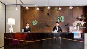 Un mostrador de recepción con dos empleados, una pared de madera con detalles decorativos y una lámpara moderna.