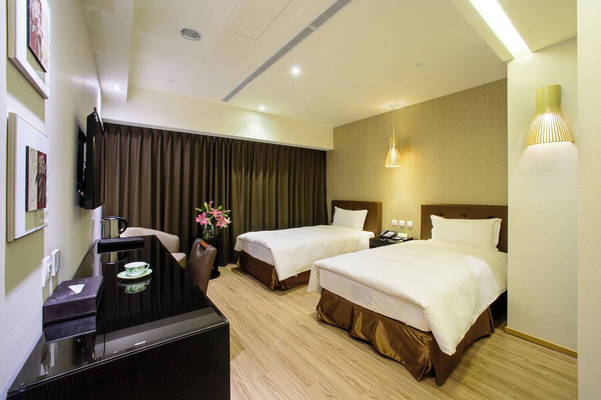 Habitación de hotel con dos camas, un escritorio con silla, televisión y una lámpara de pared.