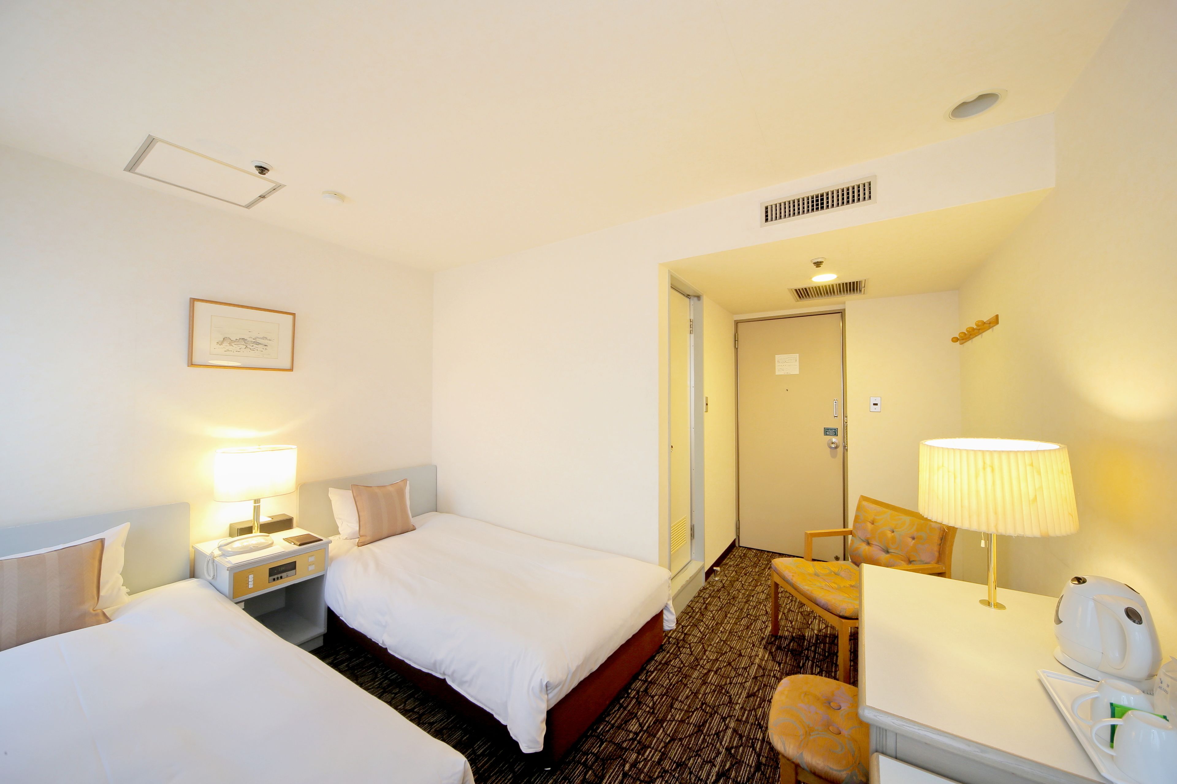 A hotel room with two beds, a desk with a lamp, a chair, a coffee maker, and a framed picture on the wall.