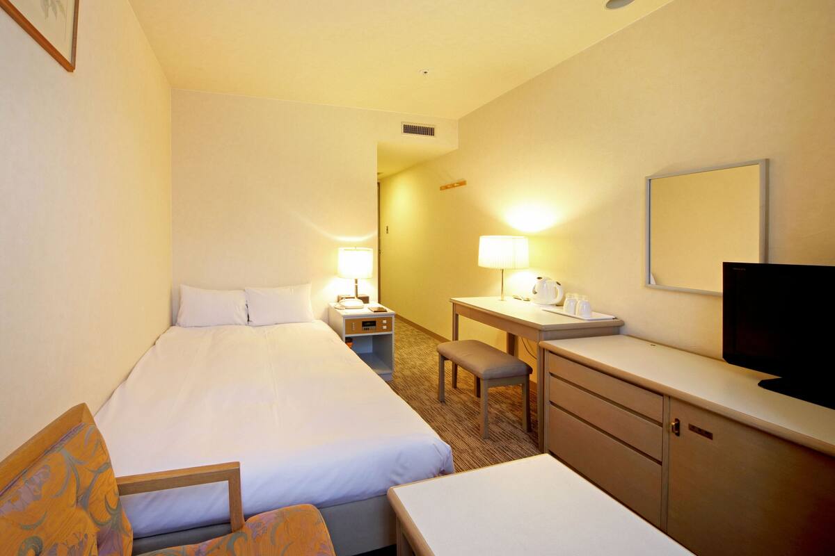 A hotel room with a bed, a desk, a chair, a television, and a nightstand with a lamp.