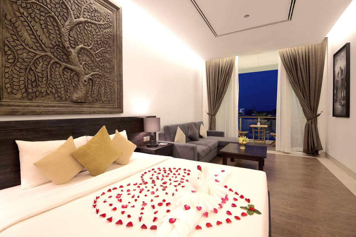Habitación de hotel con cama, sofá, mesita de noche y vistas al exterior.