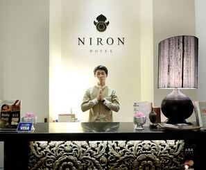 Ein Hotelrezeptionstresen mit einem Logo darüber, ein Mann steht dahinter, und es sind verschiedene Gegenstände wie eine Lampe, eine Vase und ein Kalender zu sehen.