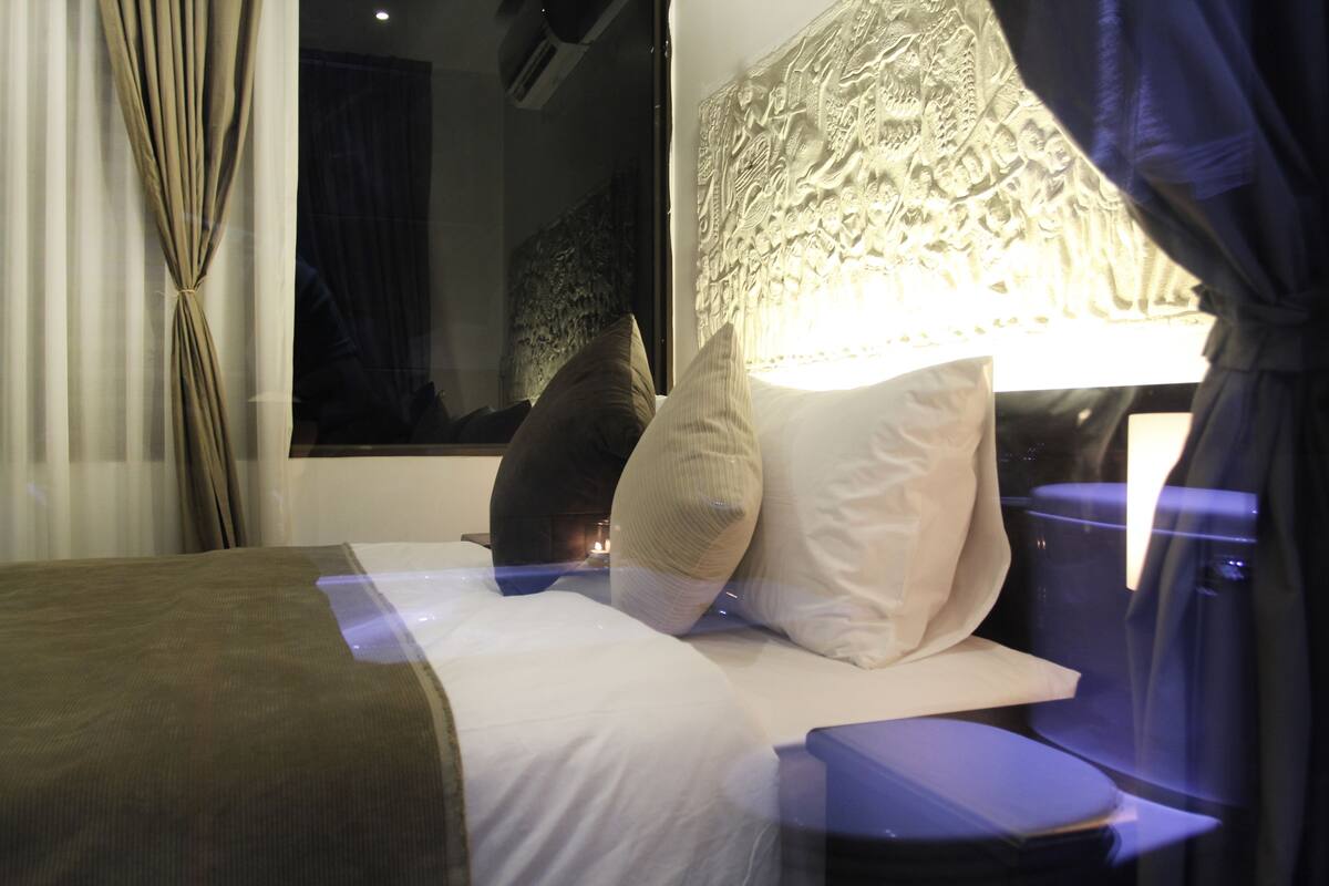 Una habitación de hotel con cama, almohadas, mesita de noche y ventana con cortinas.