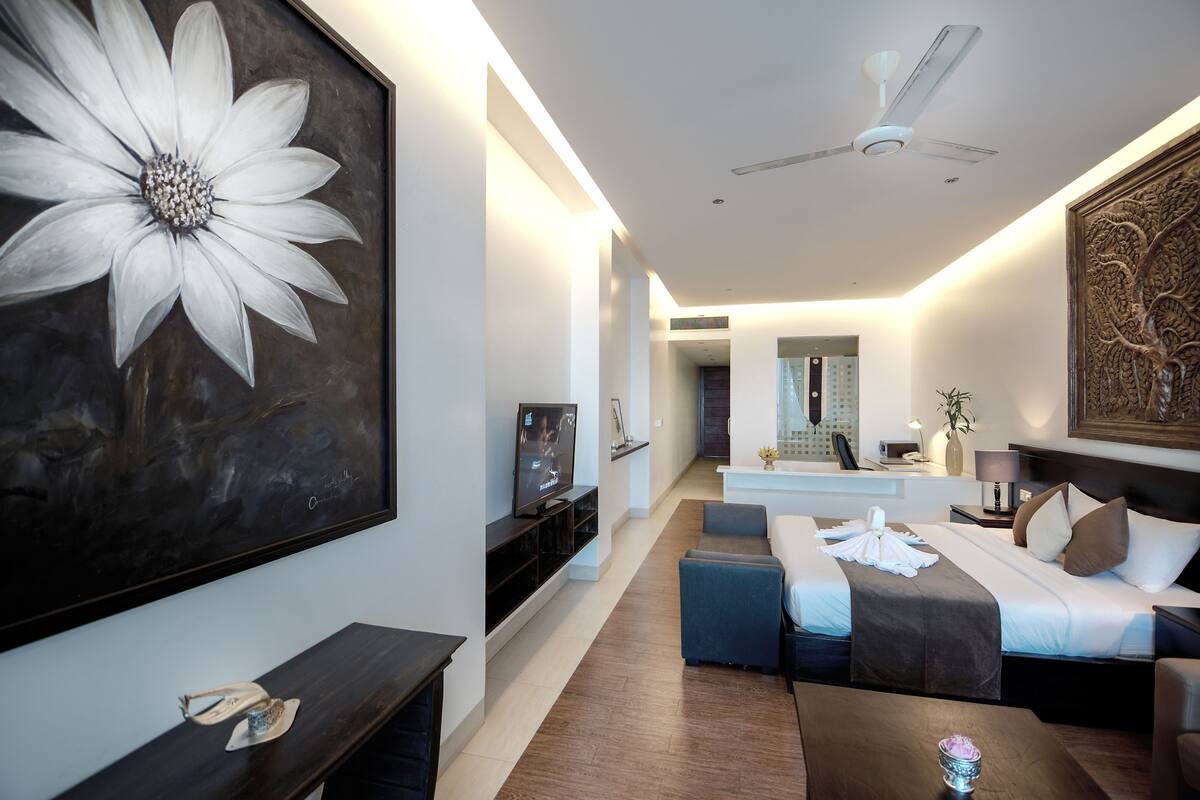 Una habitación de hotel moderna con un cuadro grande de flores, un televisor de pantalla plana, un sofá, una cama con una paleta de colores blanco y marrón, y un ventilador de techo.