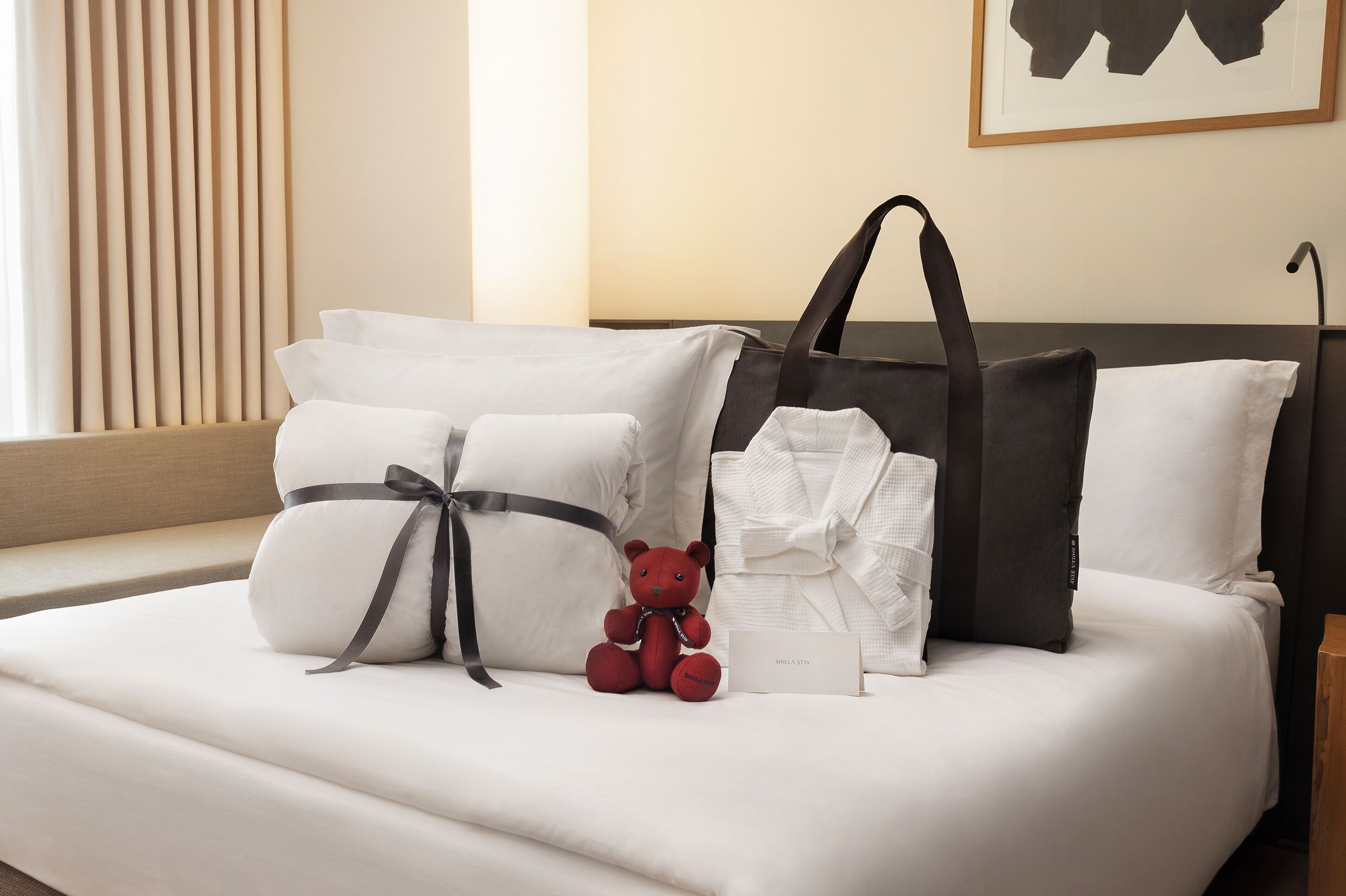 A neatly made bed with white linens, a red teddy bear, and a black and white tote bag.