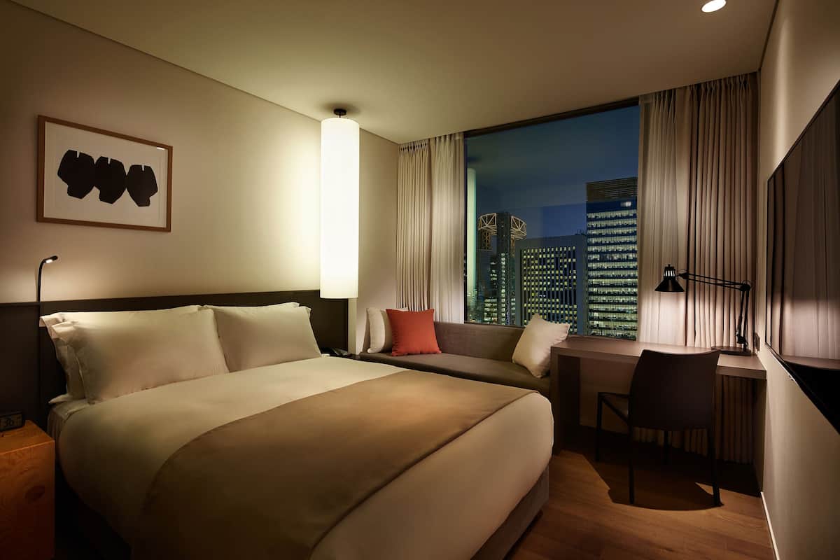 Ein Hotelzimmer mit einem großen Bett, einem Schreibtisch und Blick auf eine nächtliche Skyline.