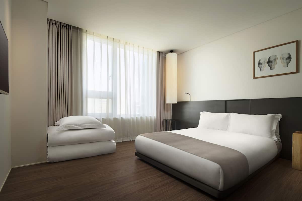 Ein modernes Hotelzimmer mit einem großen Bett, einem Nachttisch, einem Sessel und einem großen Fenster mit Vorhängen.