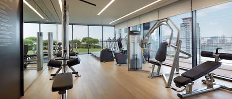 Un gimnasio moderno con diversos equipos de ejercicio, amplias ventanas y vistas a la ciudad.