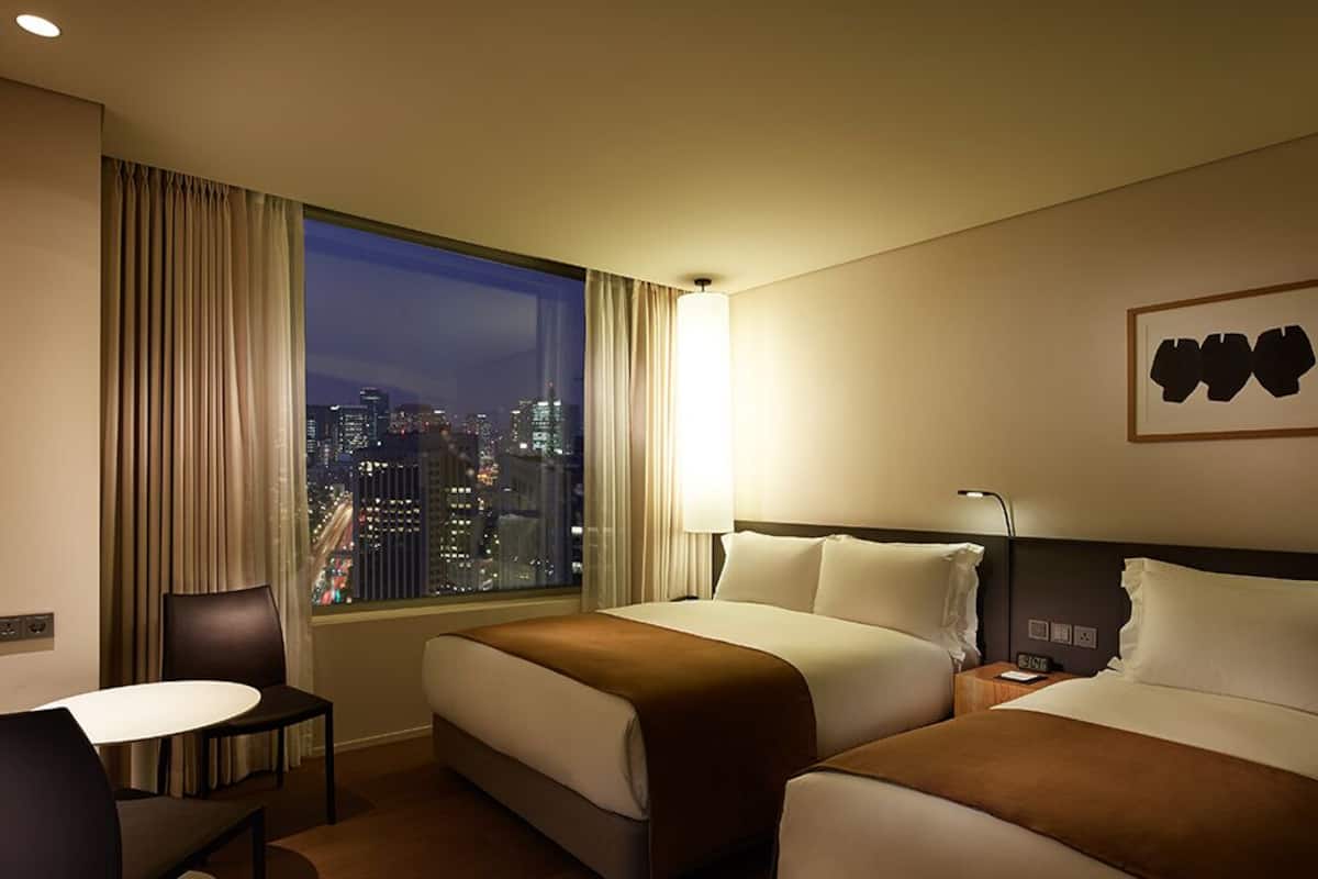 Ein Hotelzimmer mit zwei Betten, einem Schreibtisch und einem großen Fenster mit Blick auf die Stadt.