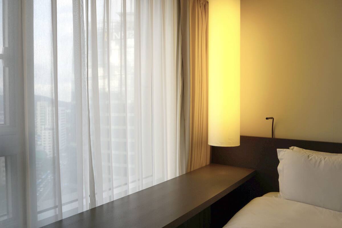 Une chambre d’hôtel avec un lit, une table de chevet, une lampe et une fenêtre avec des rideaux légers.