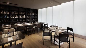 A modern dining area with multiple tables and chairs, a large bookshelf filled with decorative items, and a large window providing natural light.