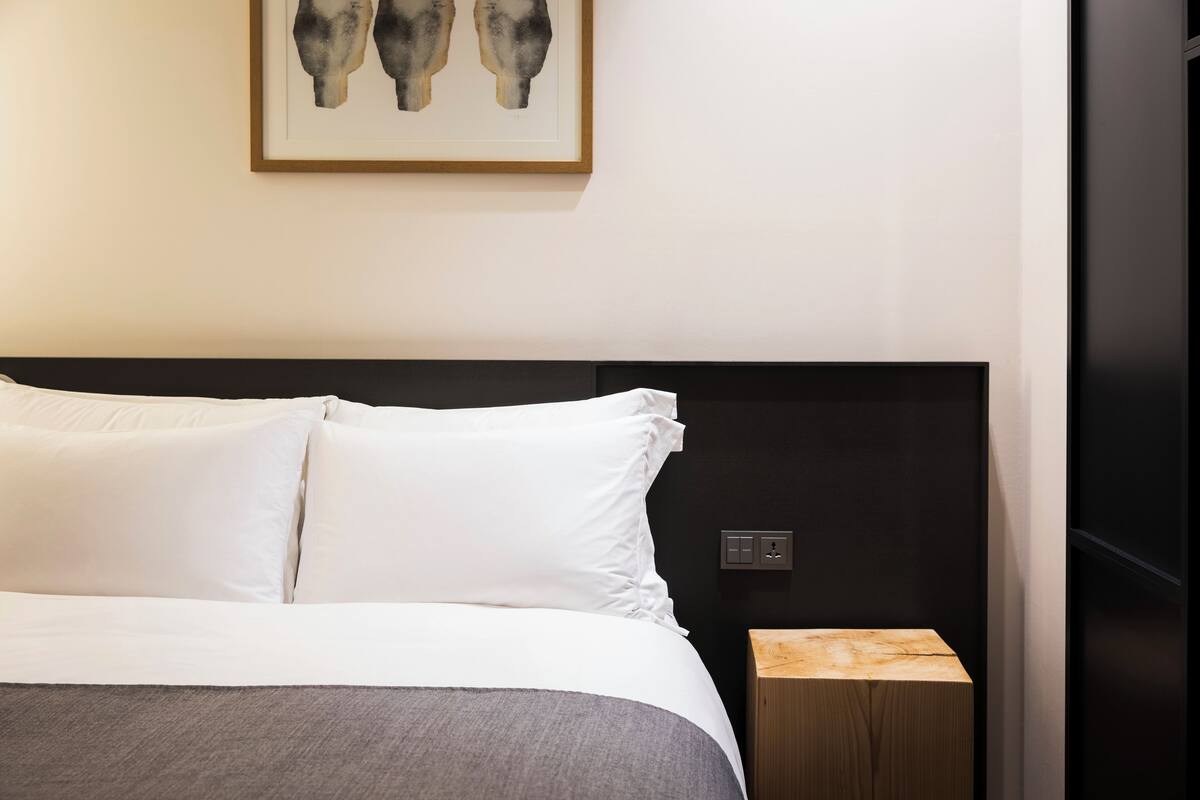 Ein ordentlich bezogenes Bett mit weißen Leinen und einer grauen Decke, ein Nachttisch daneben und ein gerahmtes abstraktes Kunstwerk an der Wand.