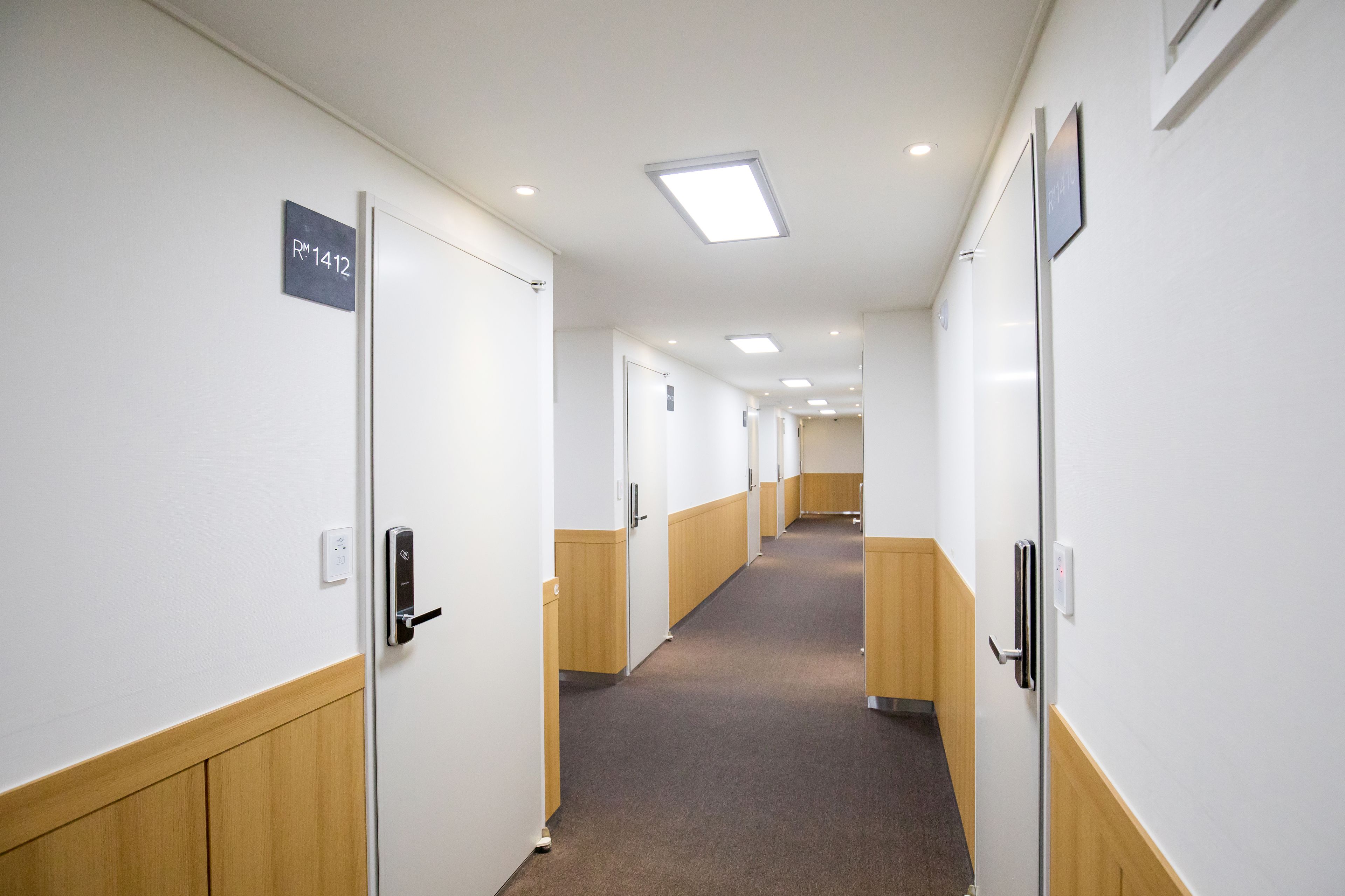 A hallway with doors marked with room numbers, wooden paneling, and recessed ceiling lights.