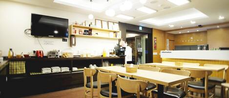 Un interior de restaurante con mesas y sillas de madera, una barra con un televisor y estantes con productos alimenticios.