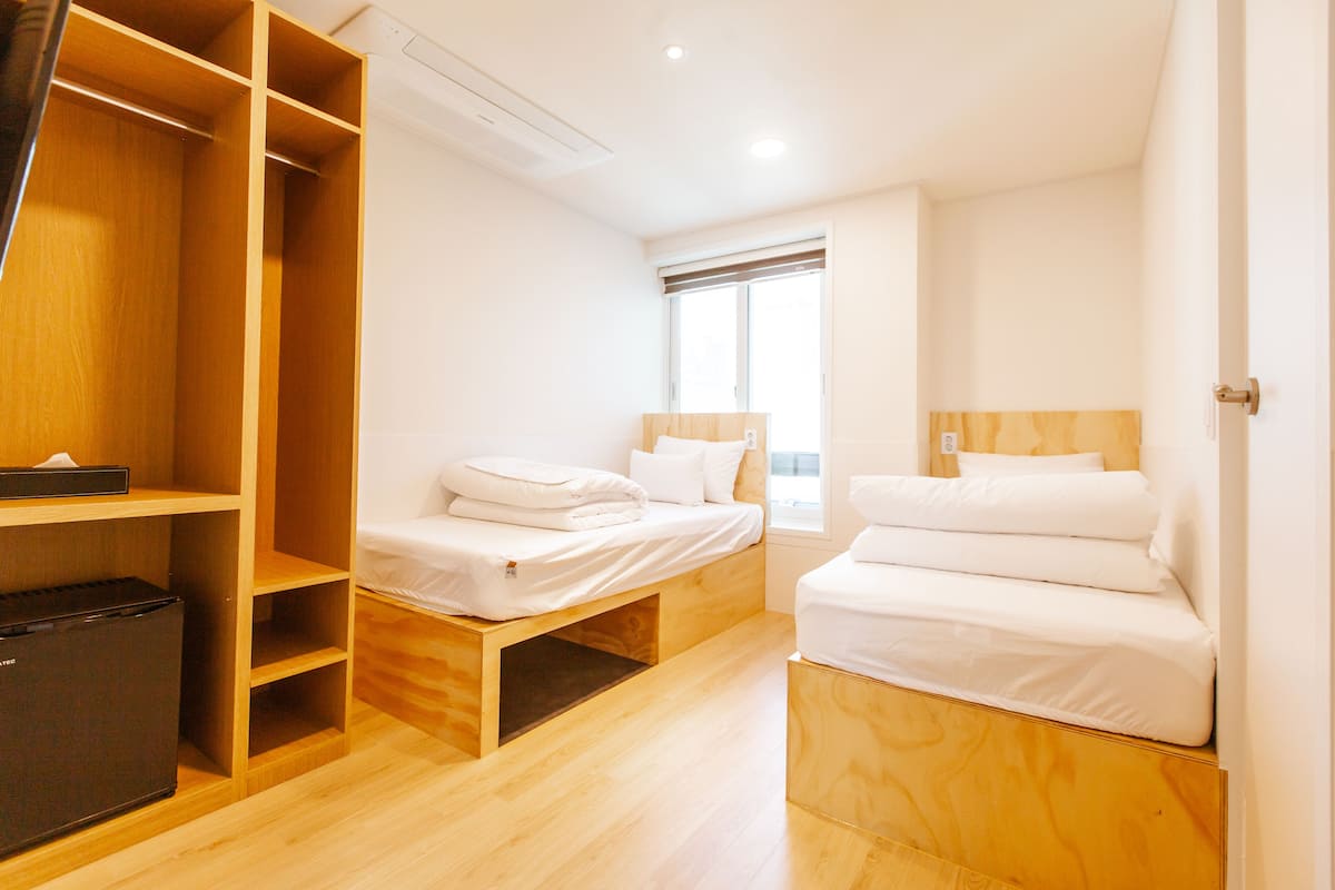 A hotel room with two beds, a wardrobe, a small refrigerator, and a window with blinds.