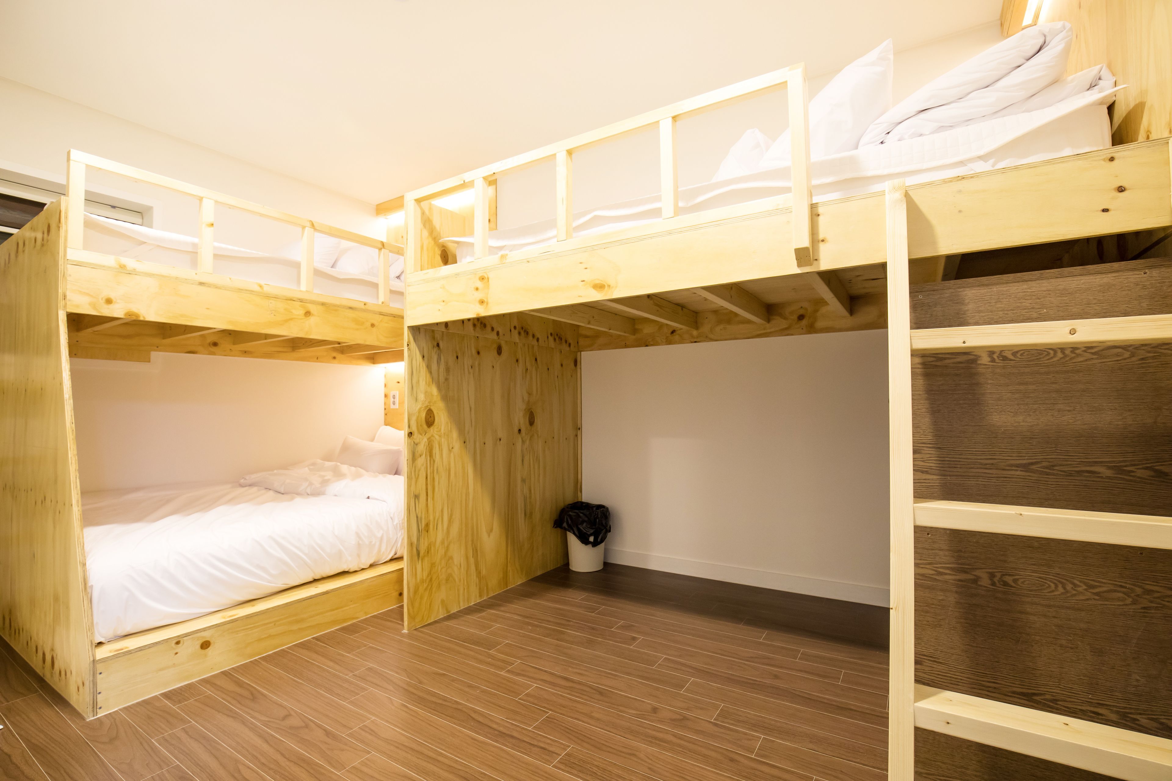 Habitación con literas de madera, un colchón blanco y un cubo de basura pequeño.