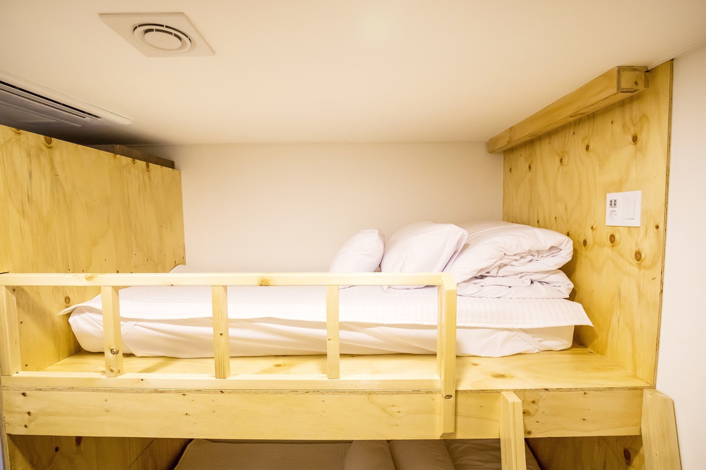 Un lit superposé avec une literie blanche, une tête de lit en bois et un mur blanc avec un interrupteur.