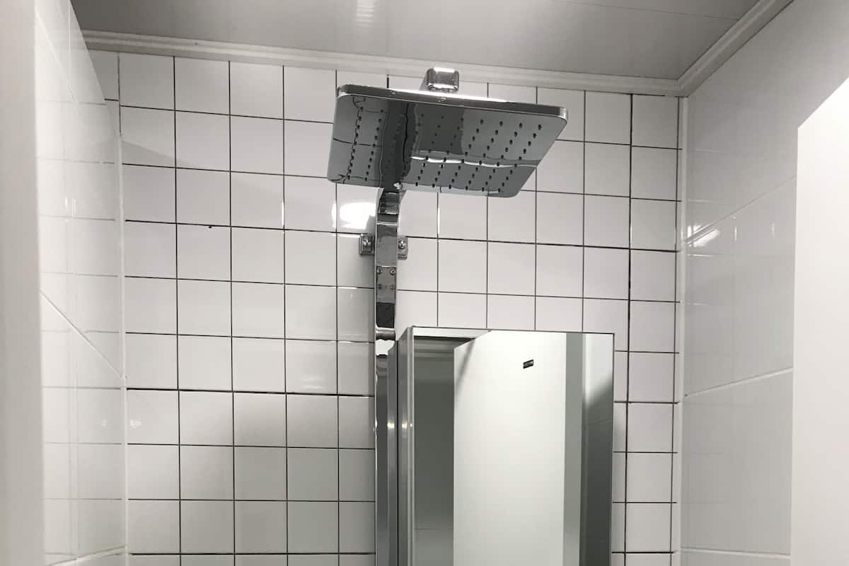 A bathroom with white tiled walls, a chrome showerhead, a mirror, and a shelf with three bottles labeled shampoo, rinse, and body wash.
