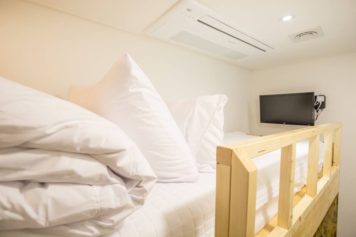 A single bed with white bedding and a wooden headboard, a television mounted on the wall, and an air conditioning unit on the ceiling.
