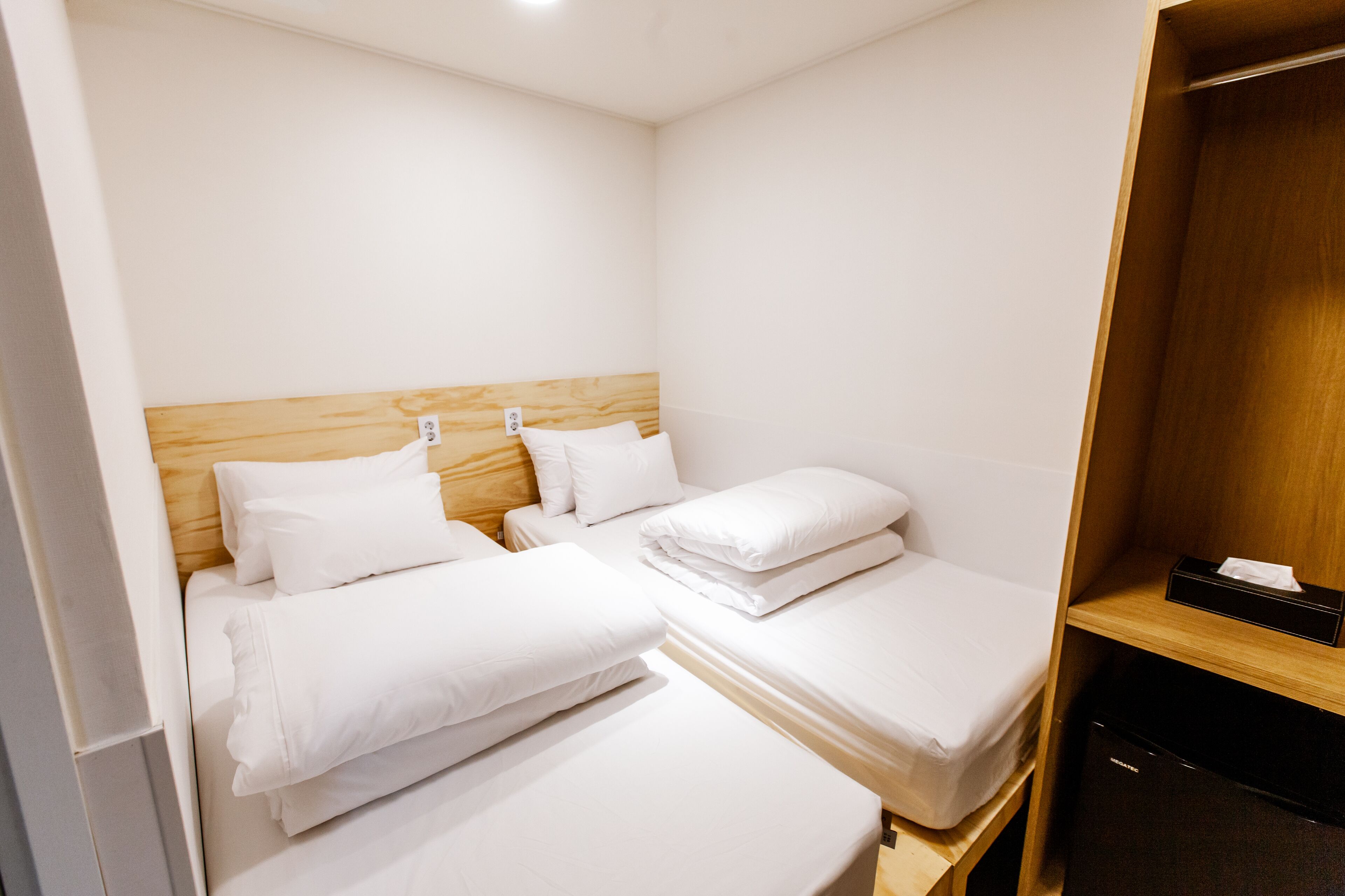 Une chambre d’hôtel avec deux lits simples, une tête de lit en bois, un mini-bar noir et une armoire en bois.