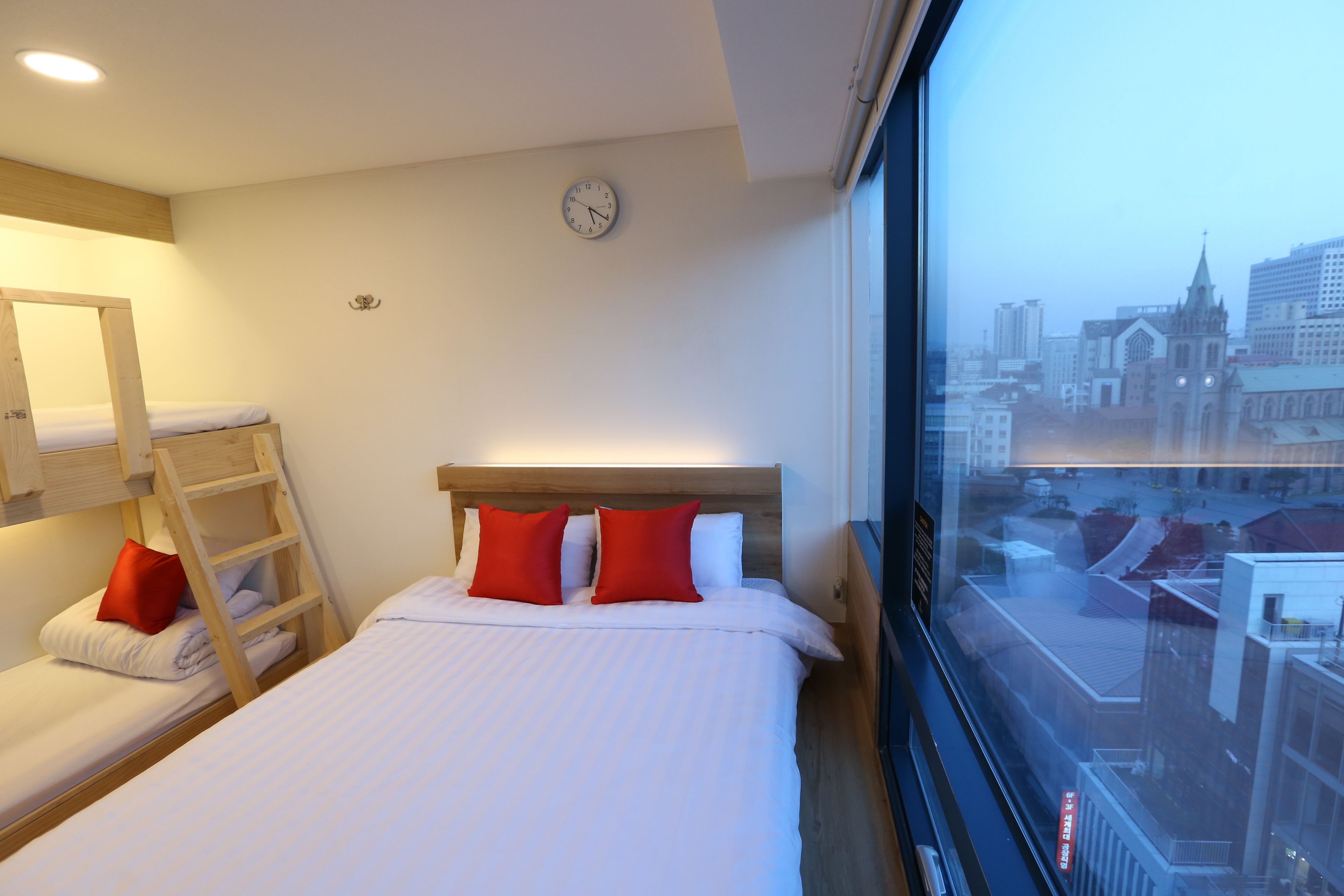 A hotel room with a bunk bed, a single bed with red pillows, a city view through a large window, and a clock on the wall.