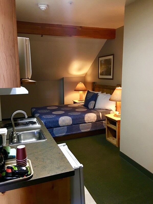 A compact hotel room with a kitchenette, a bed with a blue and white patterned bedspread, a nightstand with a lamp, and a painting on the wall.