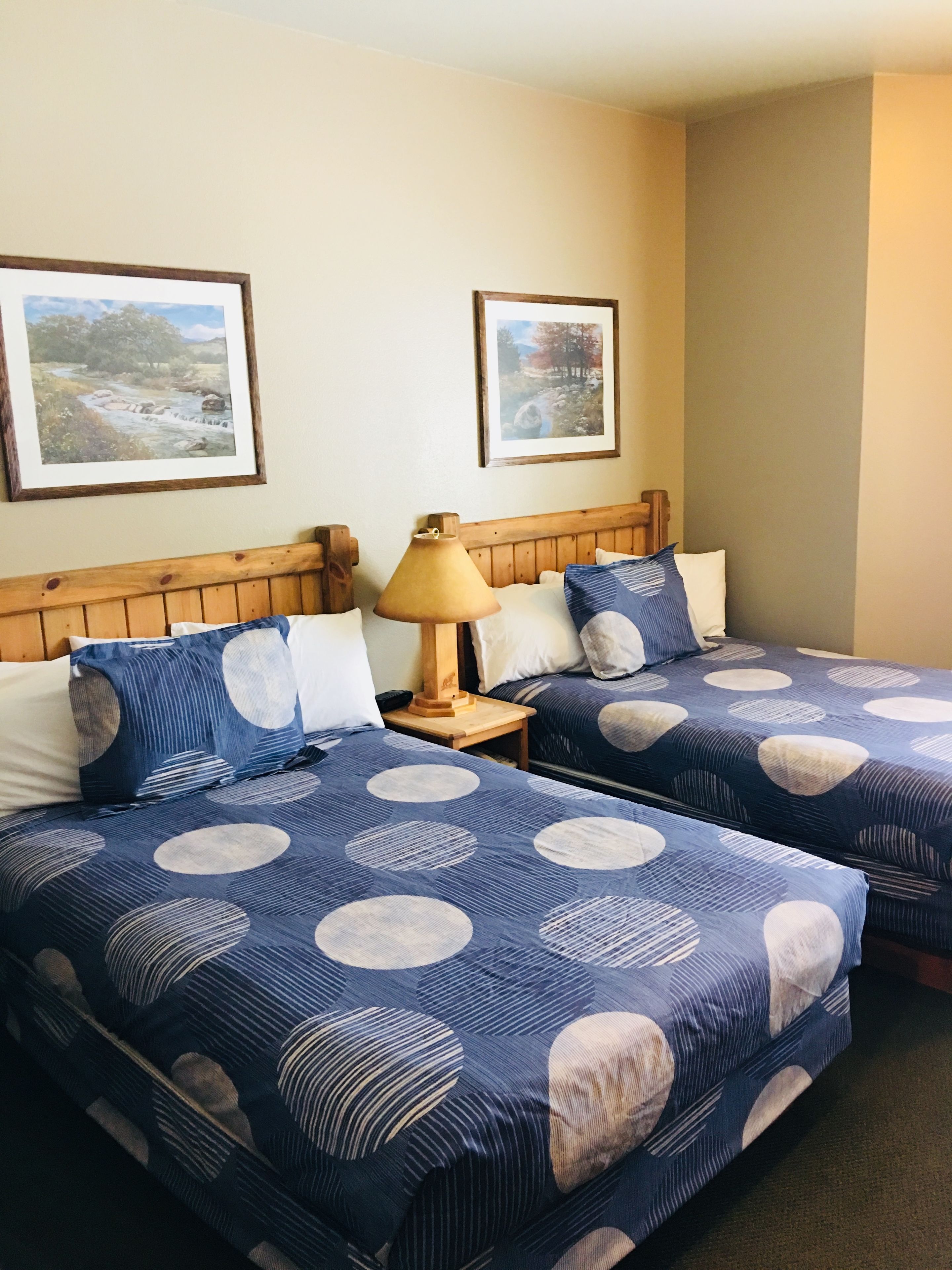 A hotel room with two beds, each with a blue and white circular pattern bedspread. There are wooden headboards, a wooden nightstand with a lamp, and two framed pictures on the wall.