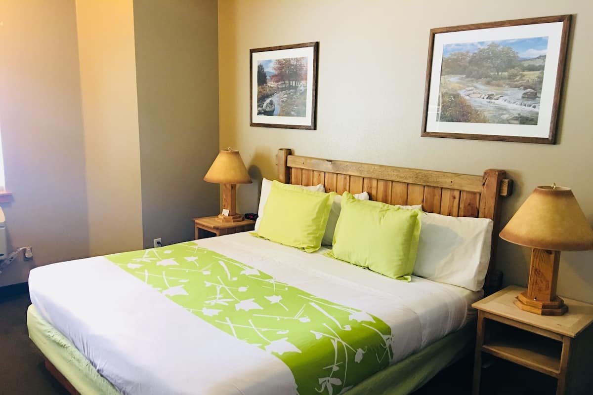 A neatly made bed with a green and white bedspread, two wooden nightstands with lamps, and two framed landscape paintings on the wall.