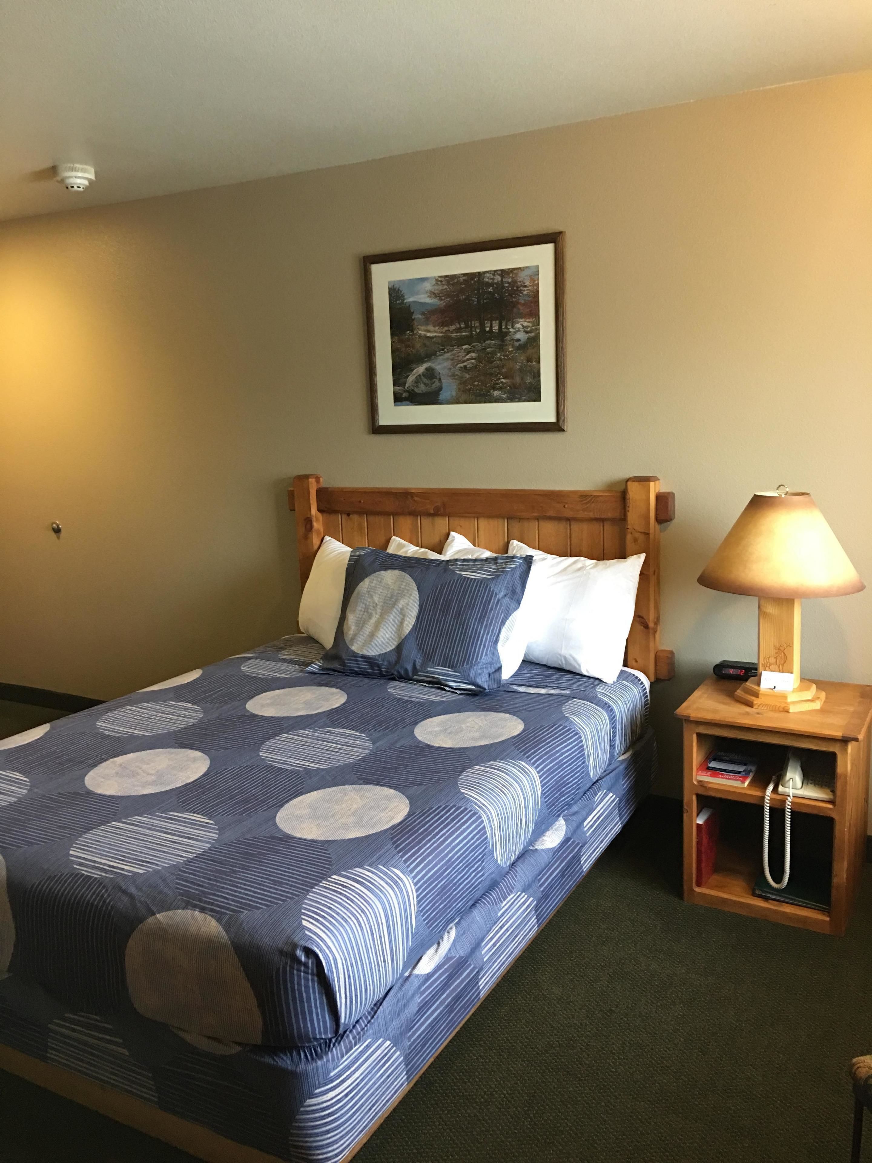 Un lit bien fait, recouvert d’une couvre-lit à rayures bleues et blanches, avec une tête de lit en bois et une table de chevet sur laquelle se trouvent une lampe et des livres.