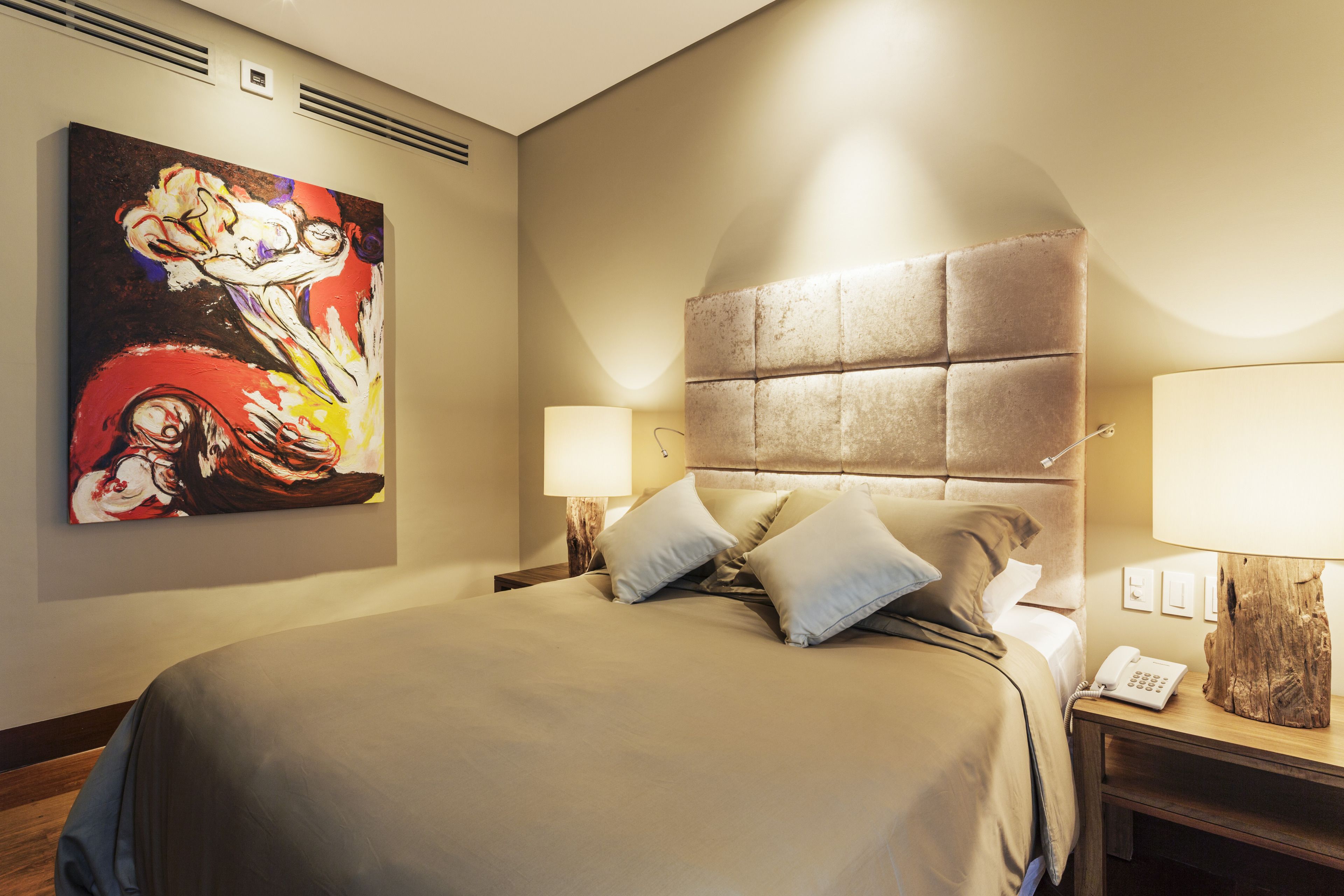 A hotel room with a large bed, a headboard, a bedside table with a lamp, a phone, and a painting on the wall.