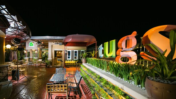 An outdoor dining area with tables and chairs, a potted plant, and a large illuminated sign. - 950 Condotel (Angeles City)
