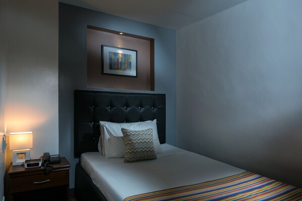 A hotel room with a bed, a nightstand with a lamp and phone, a painting on the wall, and a window.