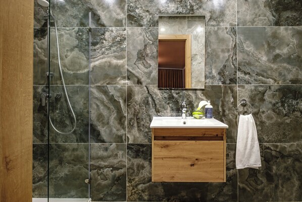A bathroom with a glass-enclosed shower, a wooden vanity with a mirror, and marble-patterned walls. - Spinola Hotel (St. Julian's)