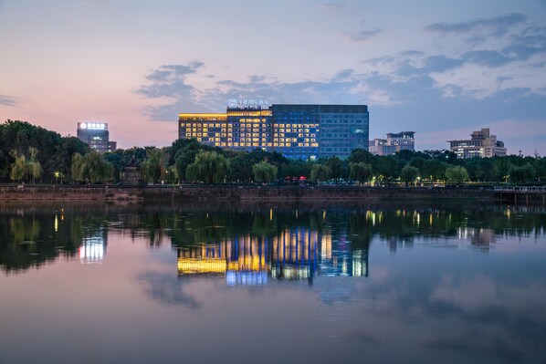 Ein modernes Hotel mit Spiegelung im Wasser, umgeben von Bäumen und wolkenlosem Himmel.