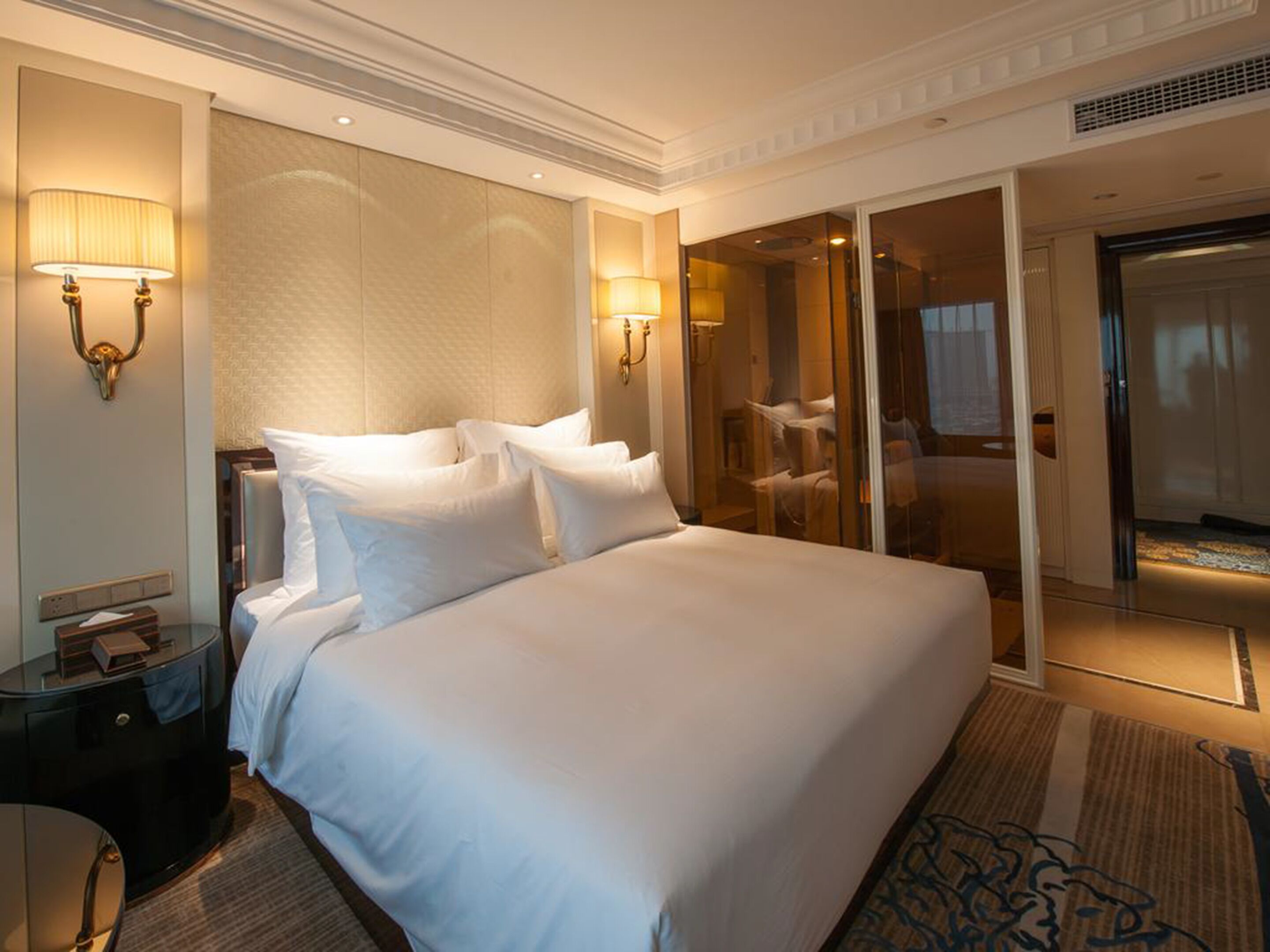 Una cama grande con sábanas blancas, dos lámparas fijadas a la pared, una puerta de cristal que da al baño y una mesita de noche con lámpara.