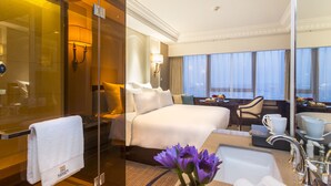 A hotel room with a bed, a desk, a chair, a bathroom with a sink, and a large window.