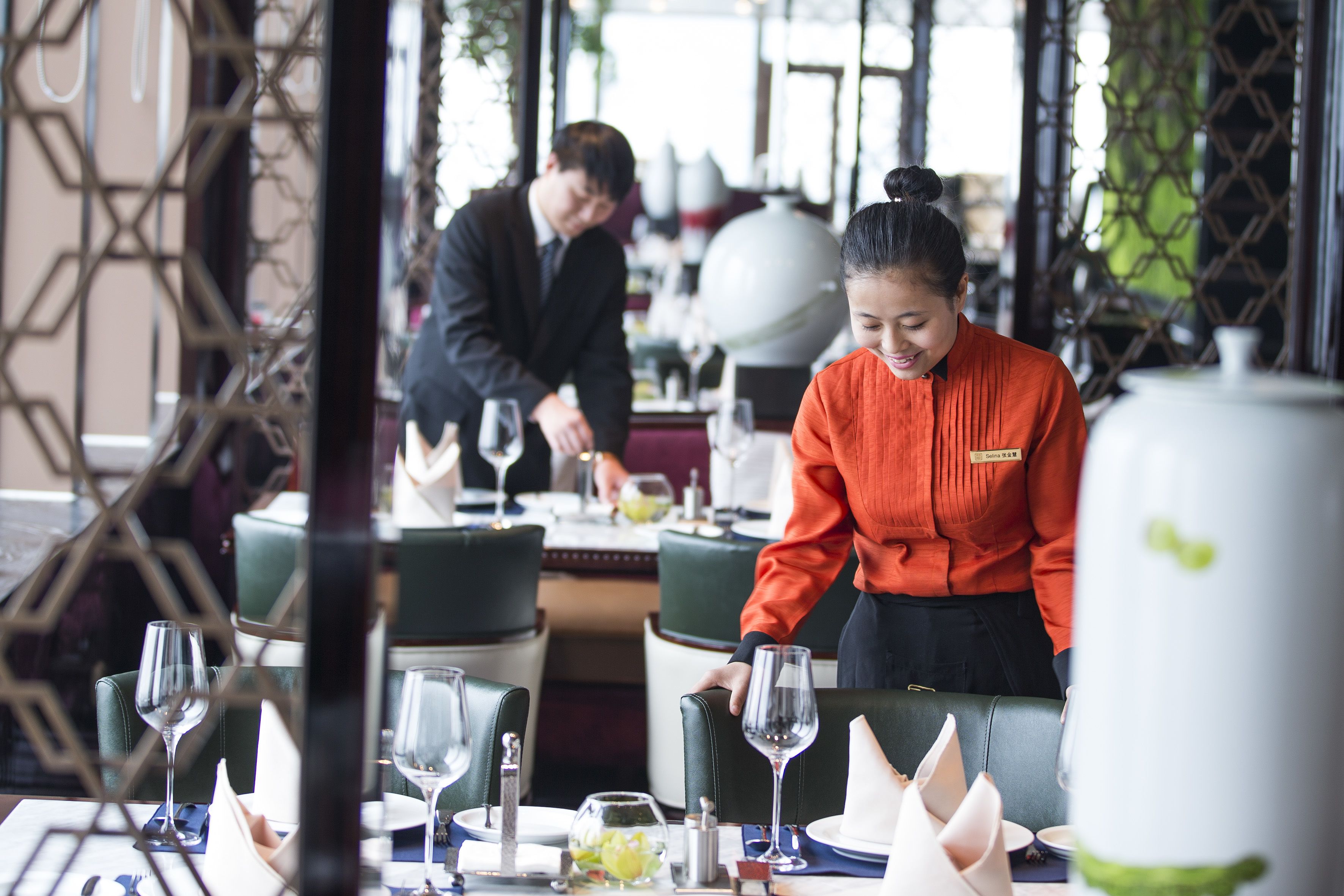 A restaurant with tables set for dining, a waiter attending to a table, and a large decorative vase on the table.