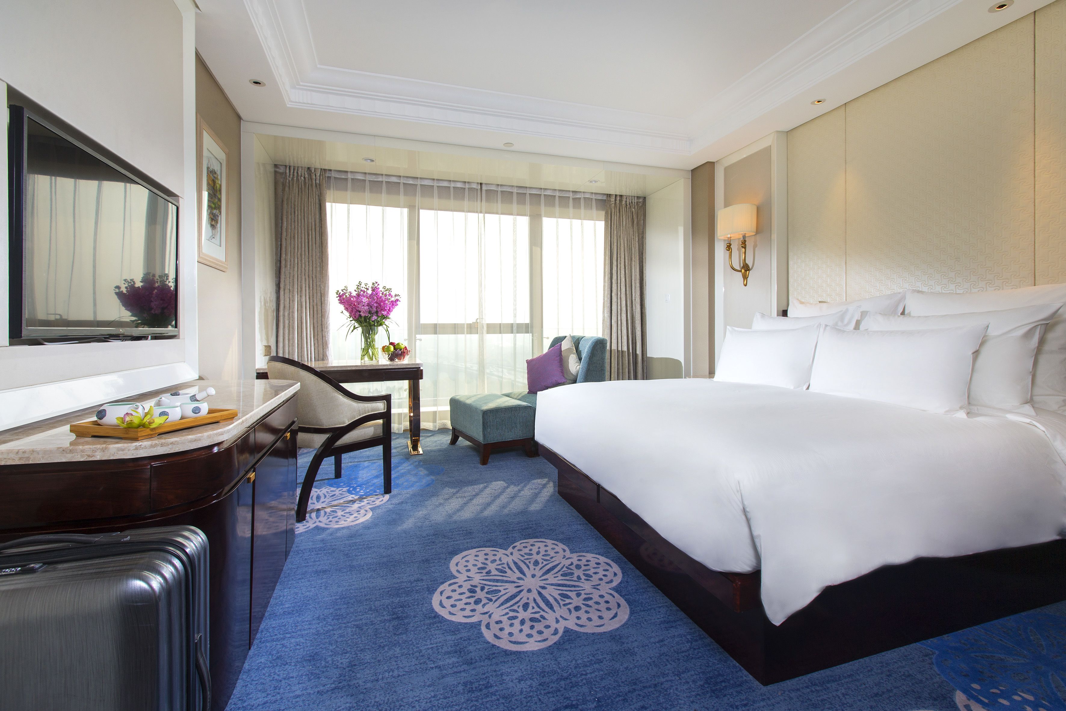 A hotel room with a large bed, a desk with a chair, a small table with a vase of flowers, and a large window with curtains.