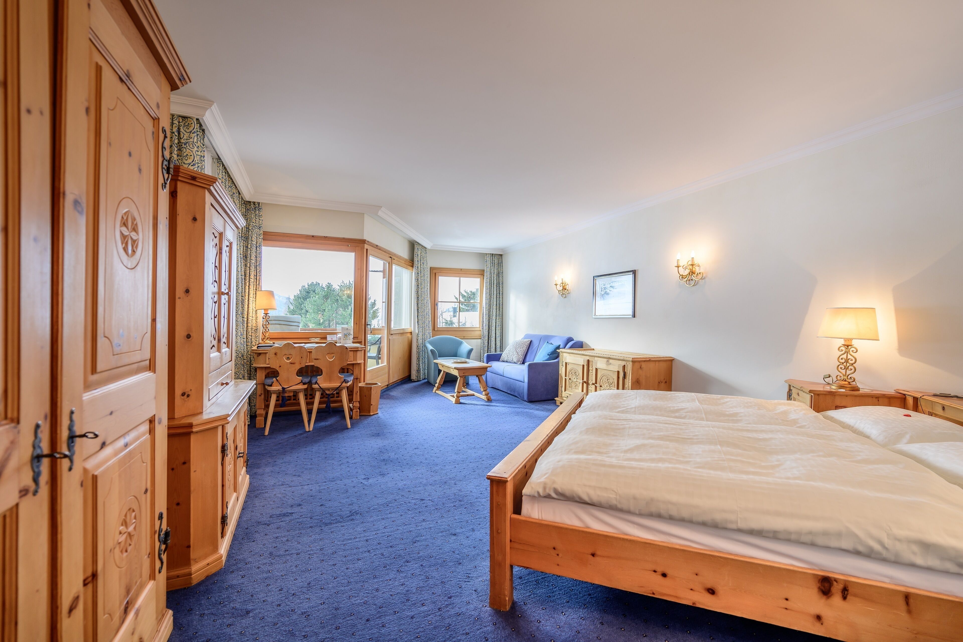 A hotel room with a large bed, wooden wardrobe, a desk with a chair, a sofa, and a view of the outdoors.