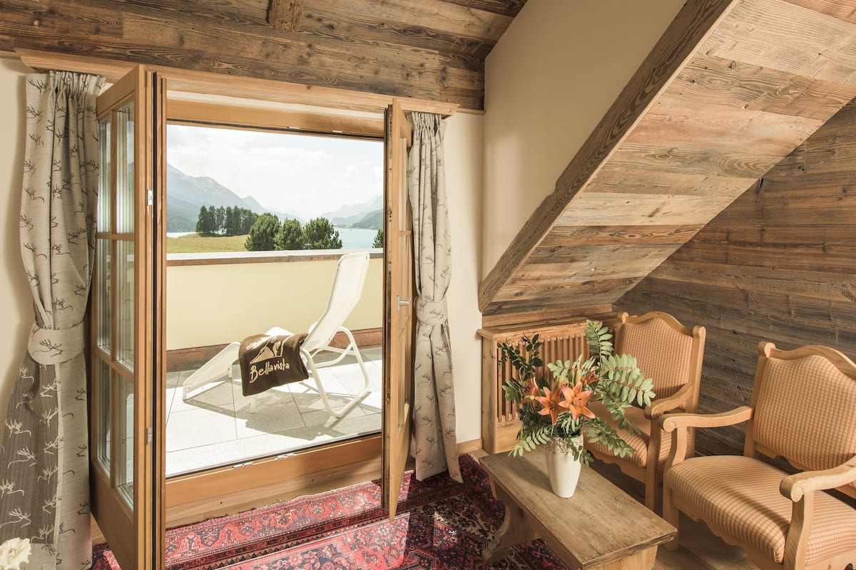 Una habitación con techo de madera, vistas a montañas, una silla, una mesa con planta en maceta y una alfombra roja estampada.