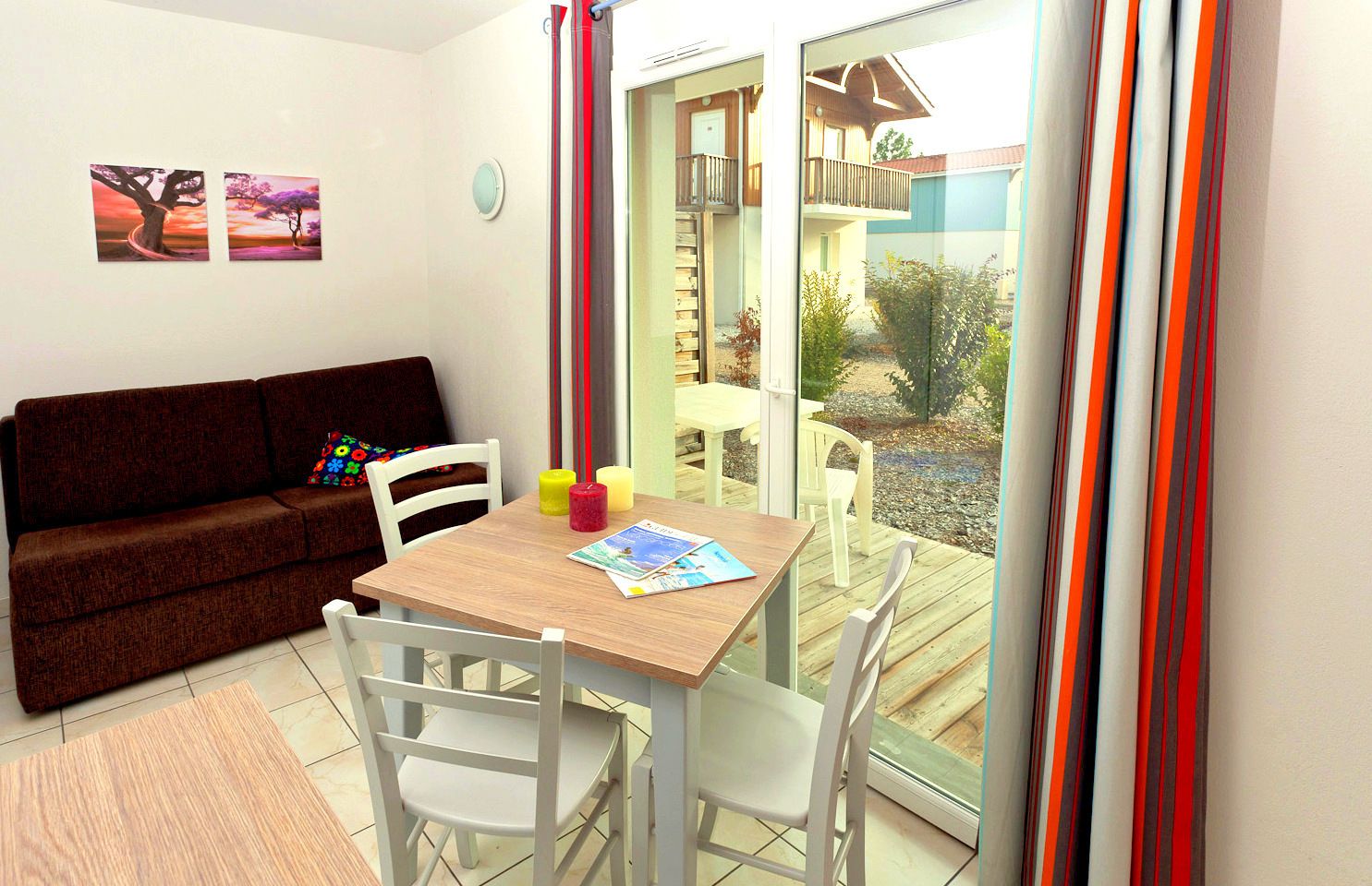 A cozy room with a brown sofa, a wooden table with chairs, and a view of a balcony with a wooden deck and plants.