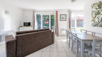 A bright living room with a sofa, a TV, a dining table with chairs, and a view of the outdoors through large windows.