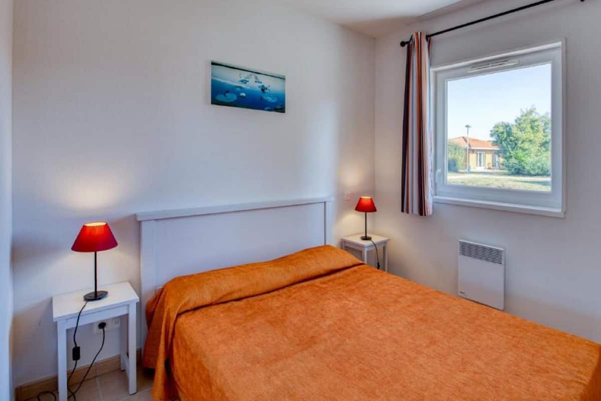 Un dormitorio con una cama, dos mesitas de noche, un ventanal con cortinas, un radiador y un cuadro colgado en la pared.