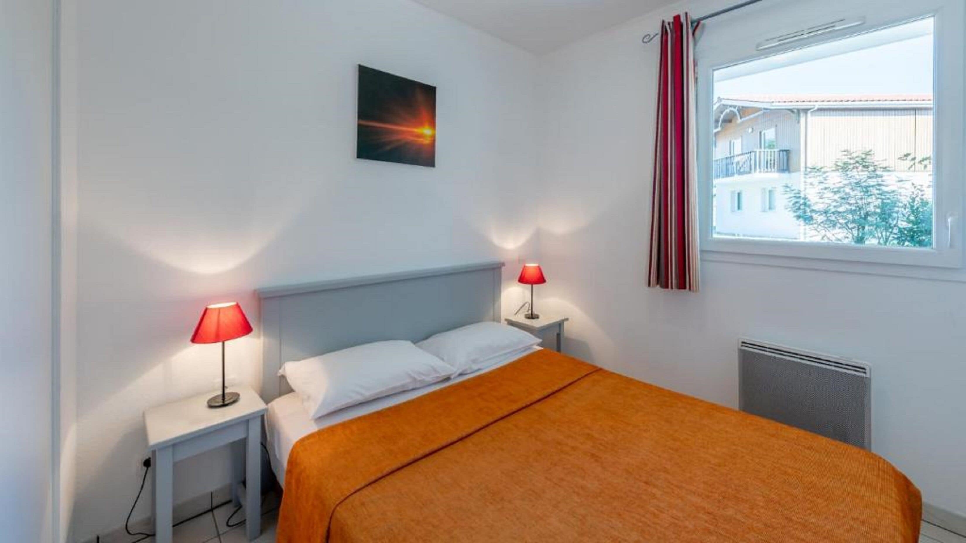 Un dormitorio con una cama, dos mesitas de noche con lámparas, una ventana con cortinas y un cuadro en la pared.