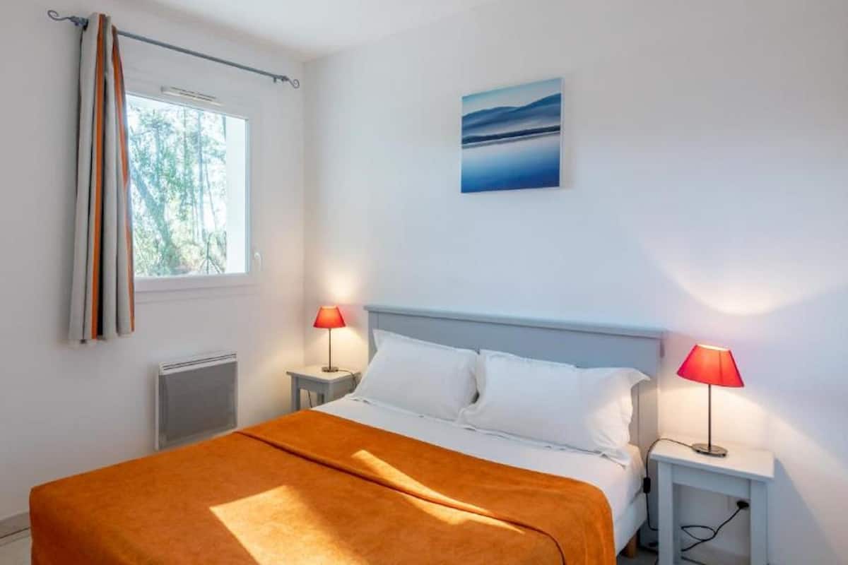 Un dormitorio con cama, dos lámparas de noche, una ventana con cortinas, un radiador y un cuadro en la pared.