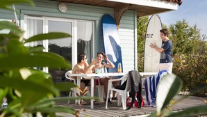 A wooden deck with a table and chairs, a surfboard leaning against a wall, and a person standing by the table.