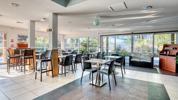 A spacious indoor area with multiple tables and chairs, large windows offering a view of the outdoors, and a seating area with a sofa and a wooden cabinet.