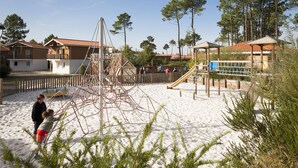 A sandy playground with climbing structures and slides, surrounded by houses with red-tiled roofs.