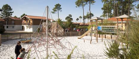 Ein Sandspielplatz mit Klettergerüsten und Rutschen, umgeben von Häusern mit roten Ziegeldächern.